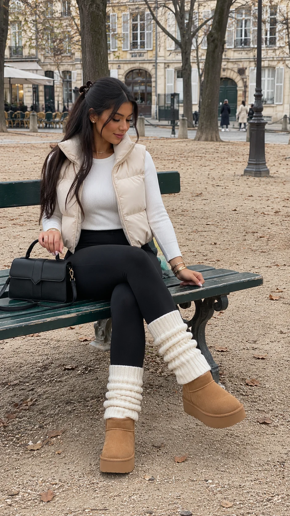 Samia assise sur un banc place Dauphine, doudoune sans manches crème, pull blanc côtelé, legging noir, guêtres en maille blanche, bottines camel à plateforme et sac noir posé à côté d'elle