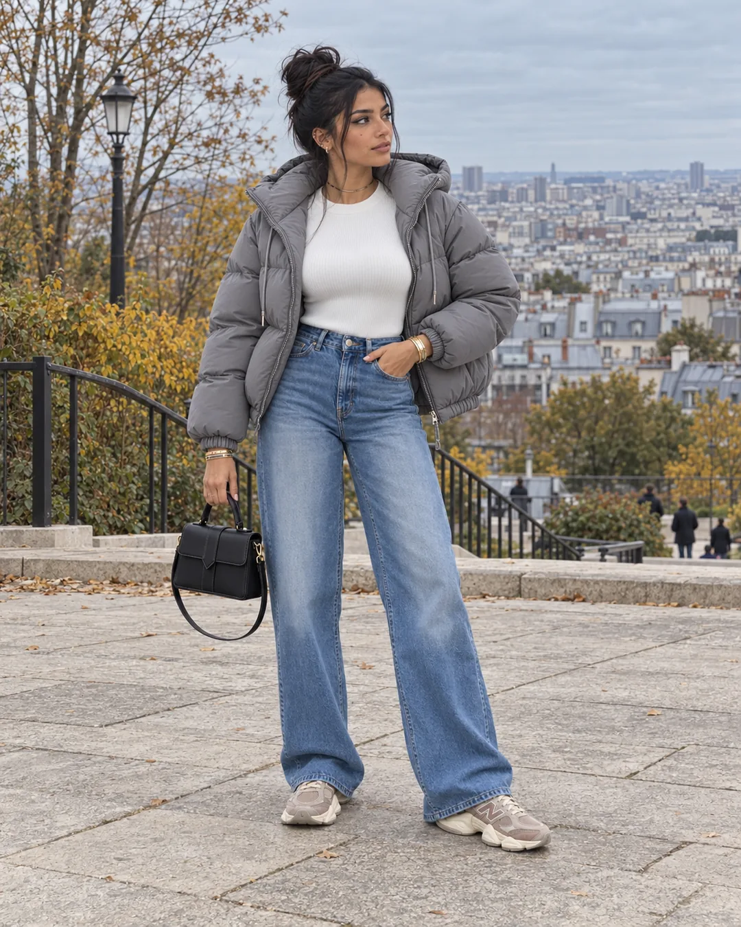 Samia debout sur la terrasse du parc de Belleville avec les toits de Paris derrière elle, doudoune grise ouverte, pull blanc côtelé, jean bleu large, petit sac noir à la main et baskets New Balance taupe